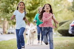 Two young girls run along a city neighborhood sidewalk, followed by their parents and dog.